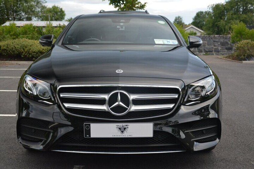 A black Mercedes-Benz car parked in a lot with a stone wall and greenery in the background. 3