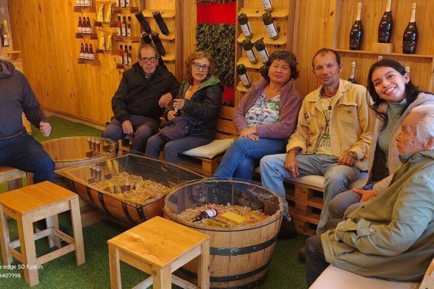 A group of people seated around a wooden table with wine bottles and barrels in a rustic setting. 5