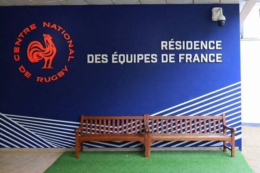 A blue wall with the text 'RÉSIDENCE DES ÉQUIPEES DE FRANCE' and a logo for 'CENTRE NATIONAL DE RUGBY'. A wooden bench is in front of the wall. 4