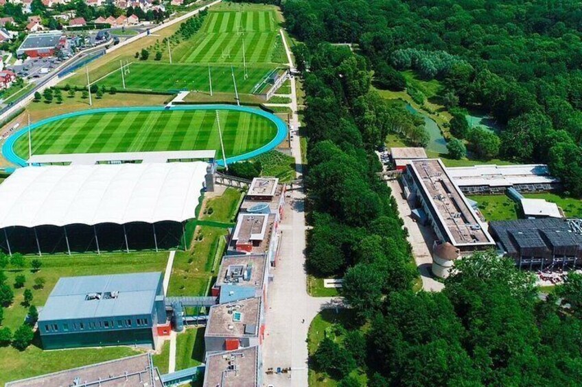 An aerial view of a sports complex with a large stadium, a running track, and surrounding greenery. 1