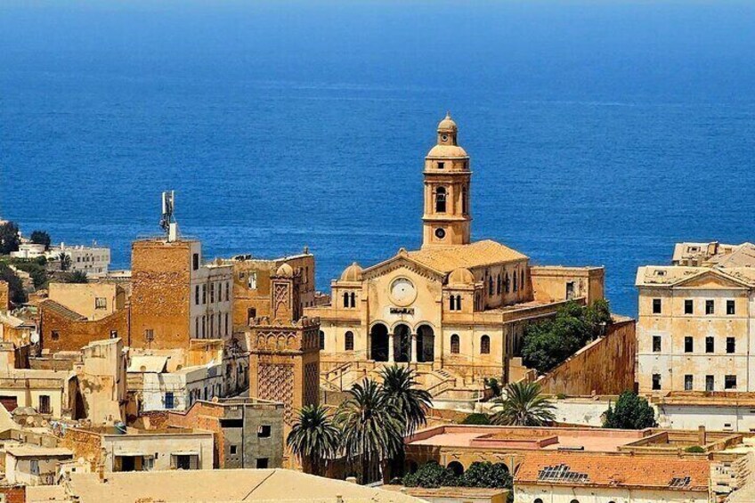 A coastal cityscape with historic architecture, including a prominent church with a bell tower, and a clear view of the sea. 3