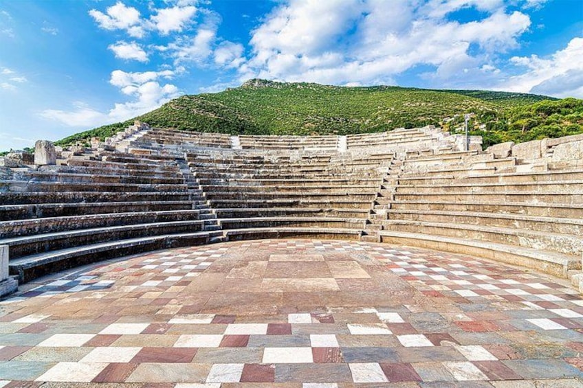 An ancient amphitheater with stone steps and a checkered floor, surrounded by a green hillside. 2