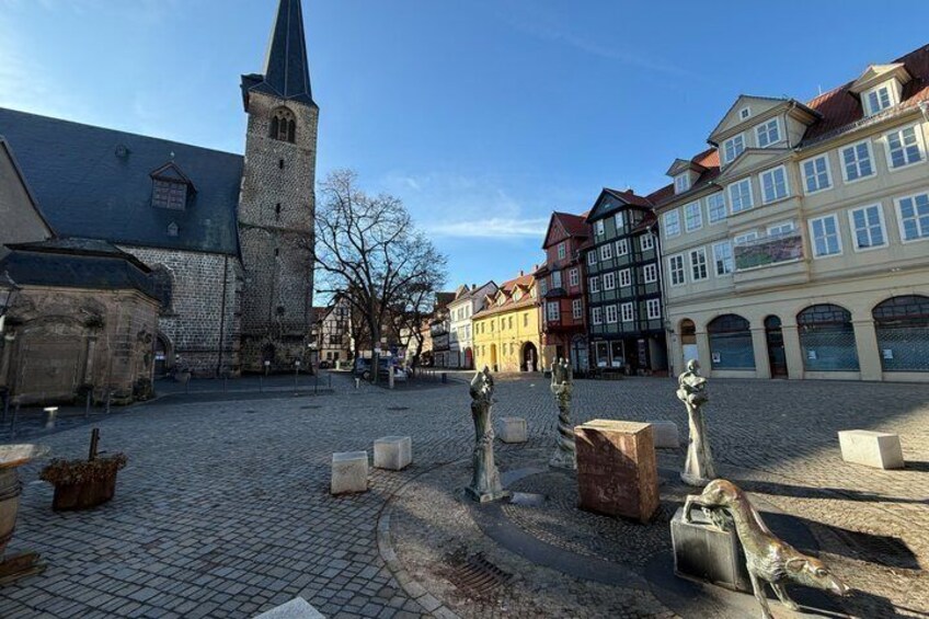 A cobblestone square with historic buildings, a church tower, and stone sculptures. 1