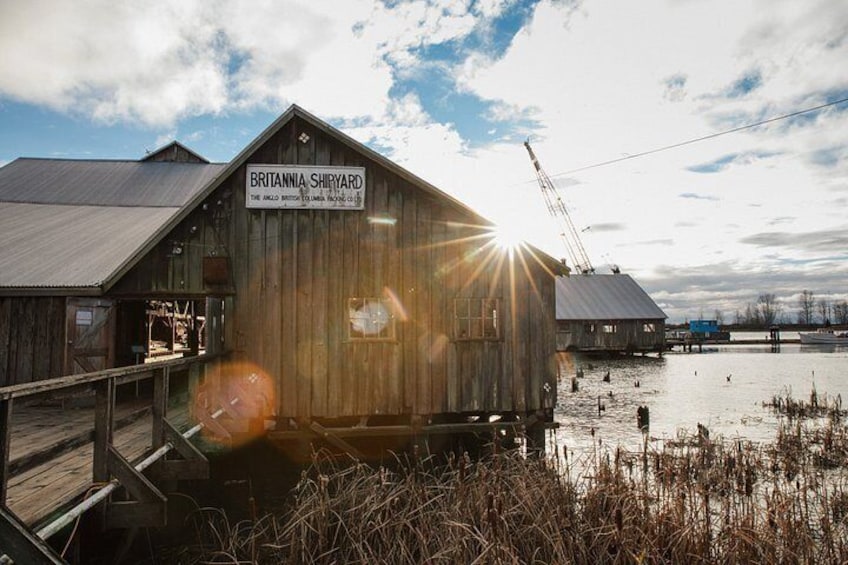 A wooden building labeled 'Britannia Shipyard' with a dock extending into a body of water, surrounded by reeds and under a partly cloudy sky. 4