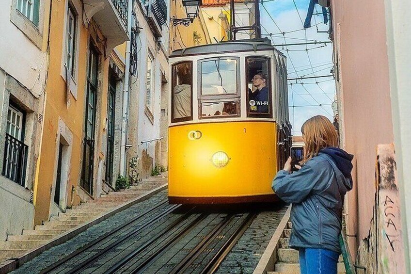 A yellow tram on tracks, a person taking a photo, and buildings on either side. 1