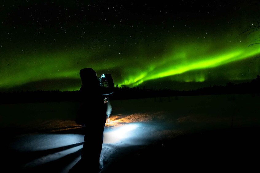 A person with a flashlight on a snowy landscape under a green aurora. 5