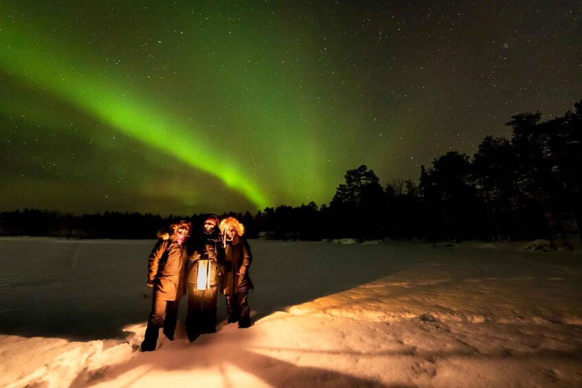 Three people standing in snow under a night sky with the aurora borealis. 1