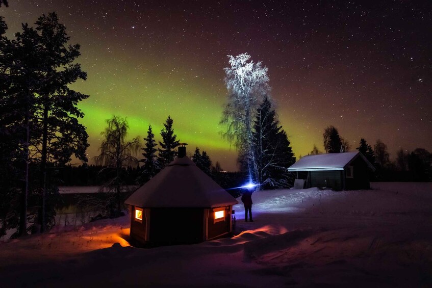 A snowy landscape with a small cabin and a larger building under a starry night sky with the aurora borealis. 2
