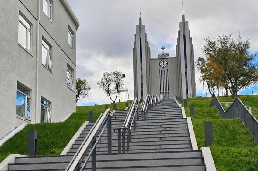 A modern building with a clock tower, a staircase leading up to it, and a grassy hill. 1