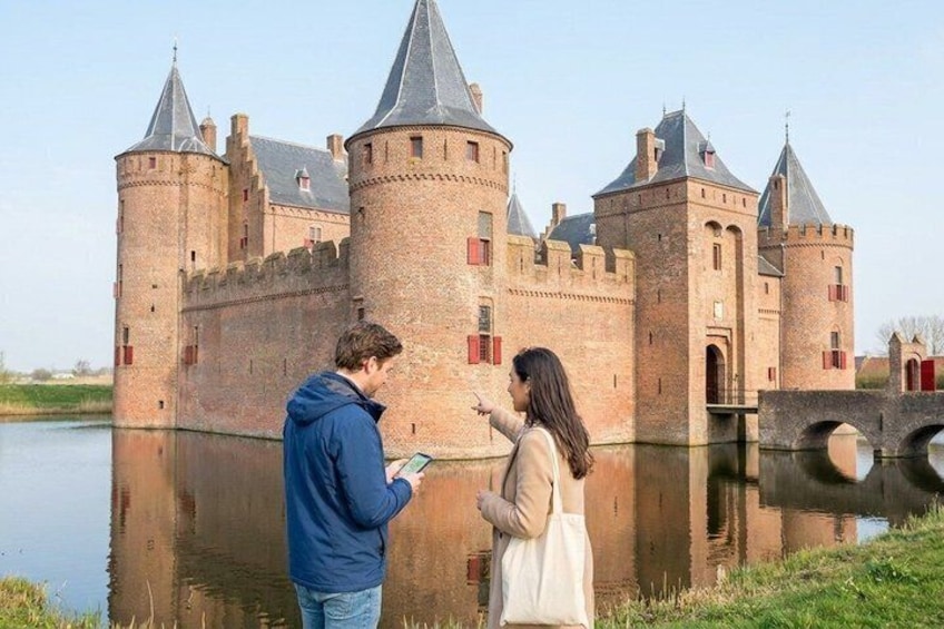 A castle by a calm river with tourists observing it. 1