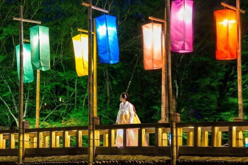 A person in traditional attire walking under colorful lanterns in a forested area. 1