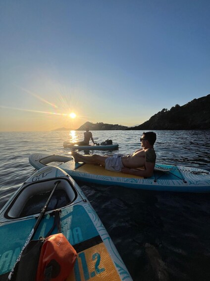 A sunset over a calm sea with a person lying on a surfboard, another person paddling a kayak, and a boat with a sail. 2