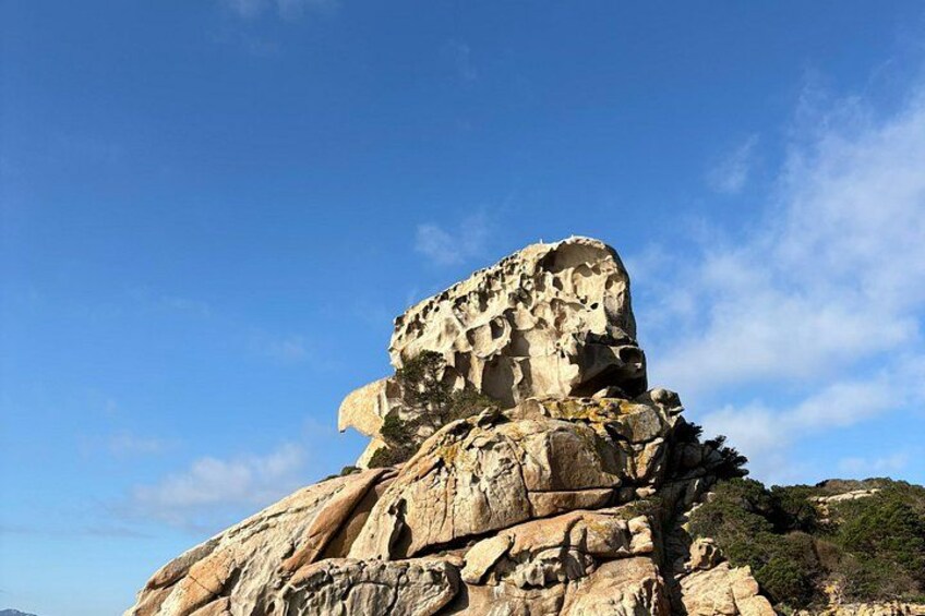 A large rock formation with a flat top and a rugged base, surrounded by clear blue sky and sparse vegetation. 3