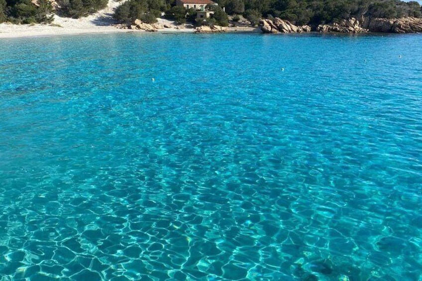 Clear turquoise sea, sandy beach, rocky coastline, and a building in the background. 4