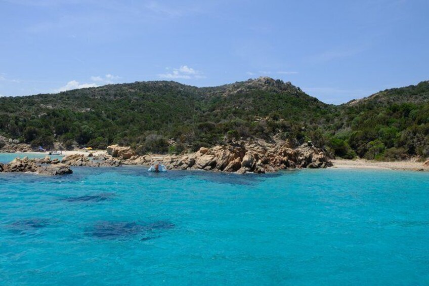 A coastal landscape with clear turquoise waters, rocky outcrops, and a hilly backdrop. 2