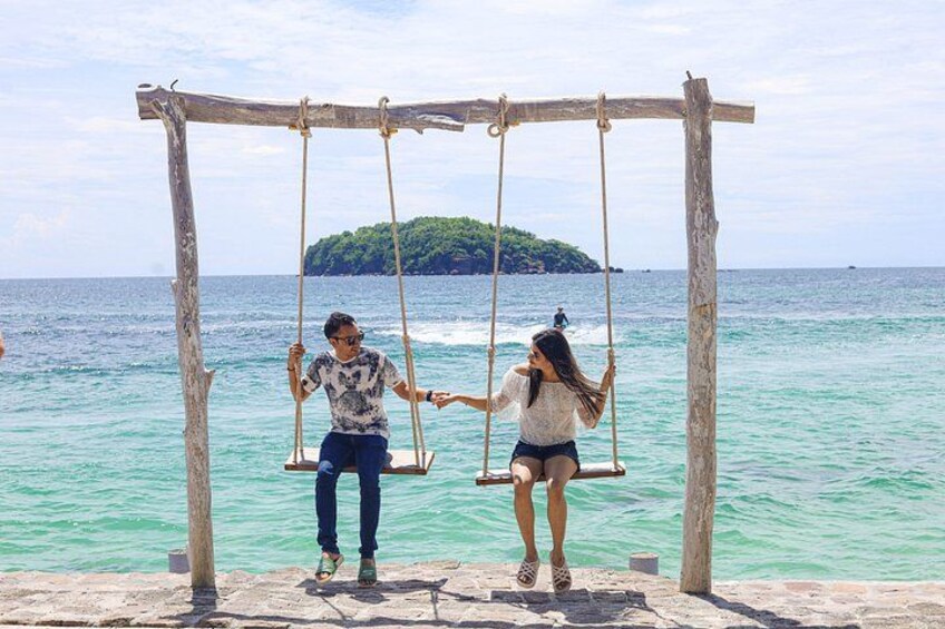 Two people on a wooden swing set over turquoise water with a small island in the background. 5