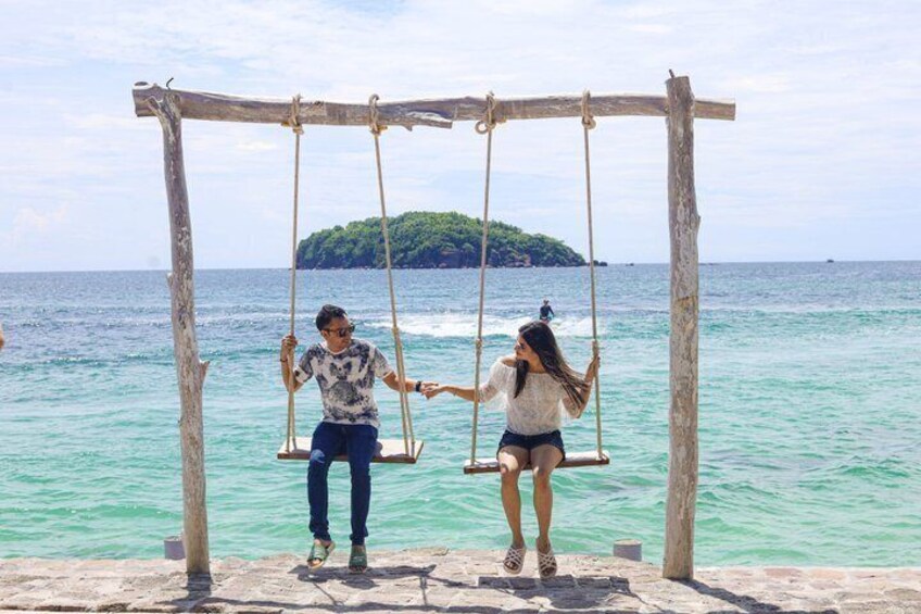 Two people on a wooden swing set over turquoise water with a small island in the background. 5