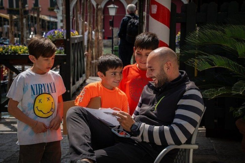 A man sitting on a bench with two children standing beside him, in an outdoor urban setting with buildings and plants in the background. 5