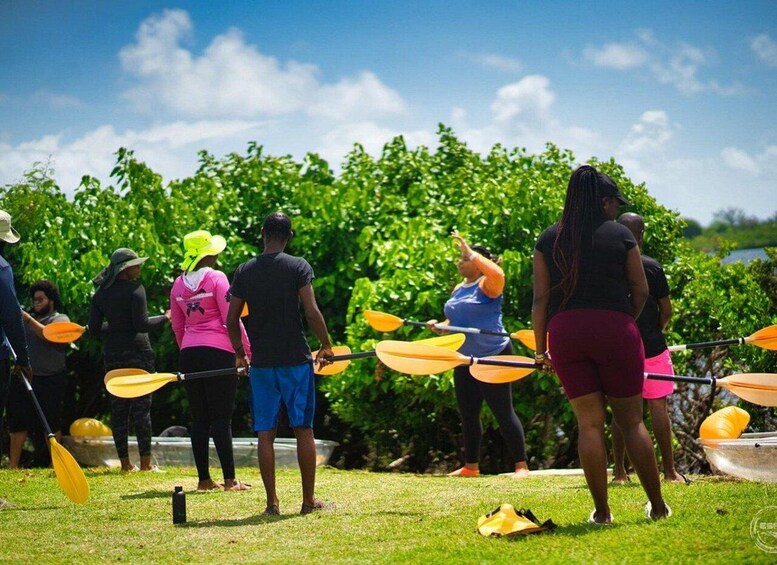 A group of people standing on grass, holding paddles, and facing a body of water. 1