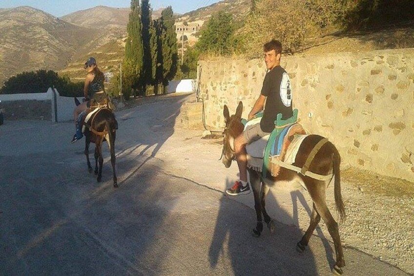 Two people riding donkeys on a paved road with a stone wall and mountains in the background. 2