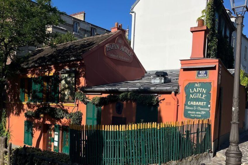 A building with a sign reading 'Au Lapin Agile Cabaret' and a green fence. 2