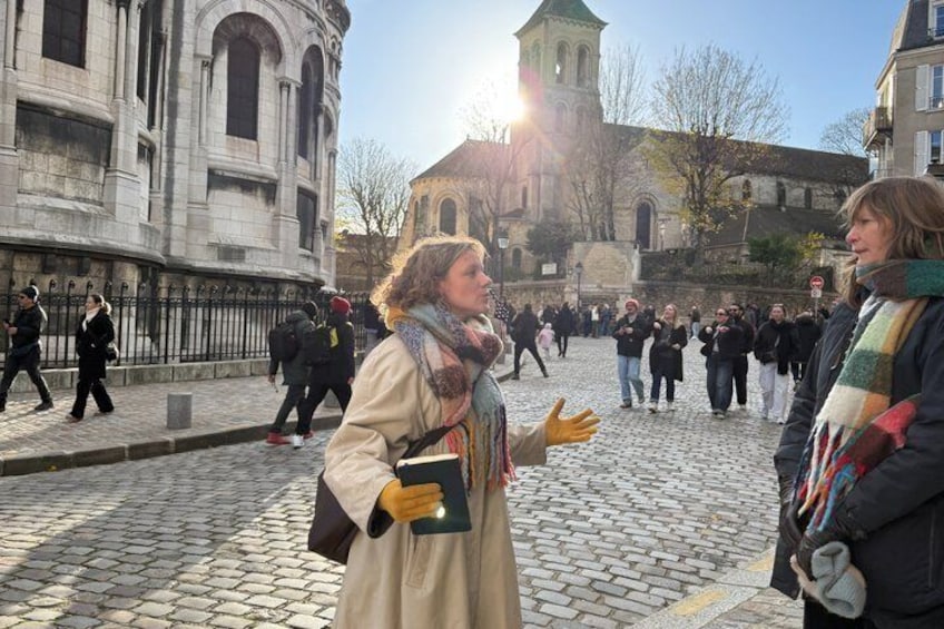 A cobblestone street with pedestrians, historic buildings, and a church tower in the background. 1