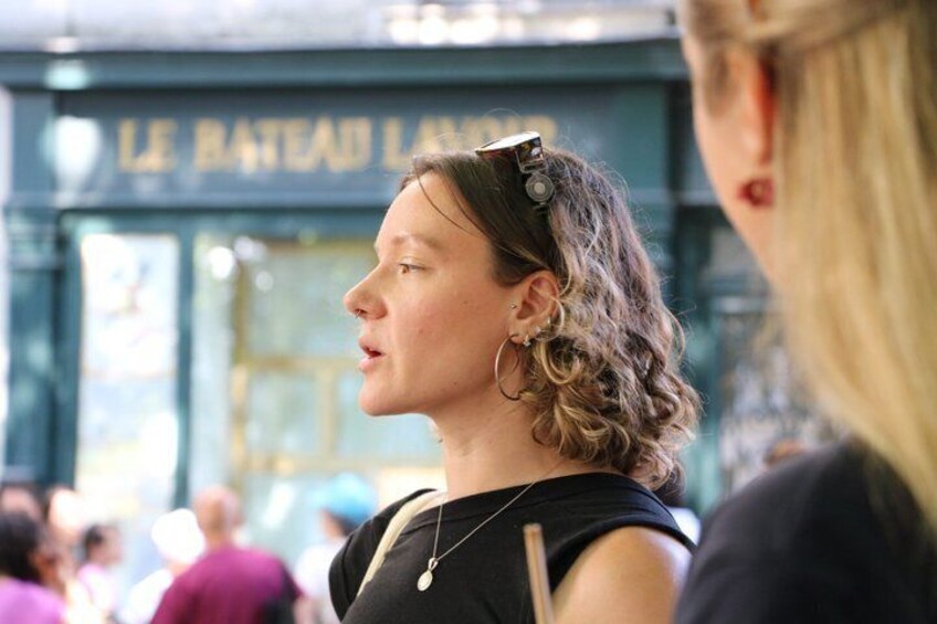 A person with curly hair wearing a black top and a necklace, standing in front of a building with a sign that reads 'LE BATEAU L'AMOUR'. 3