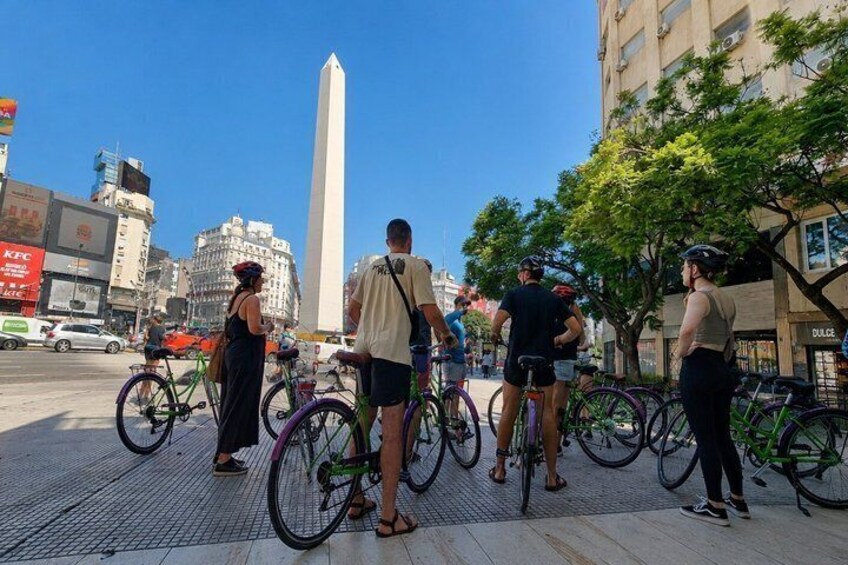A group of people standing on a sidewalk with bicycles, a tall monument, and buildings in the background. 4