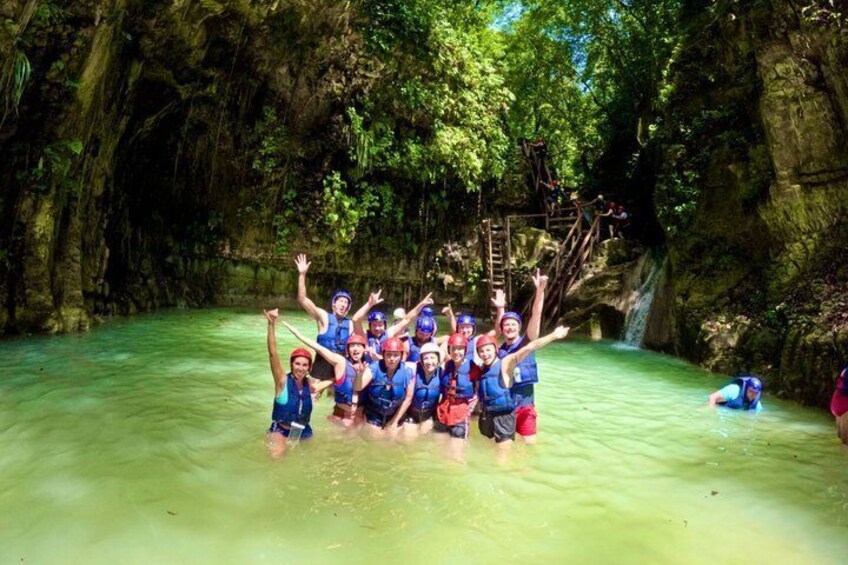 A group of people in blue life vests posing in a clear, greenish water body surrounded by rocky cliffs and dense greenery. 1
