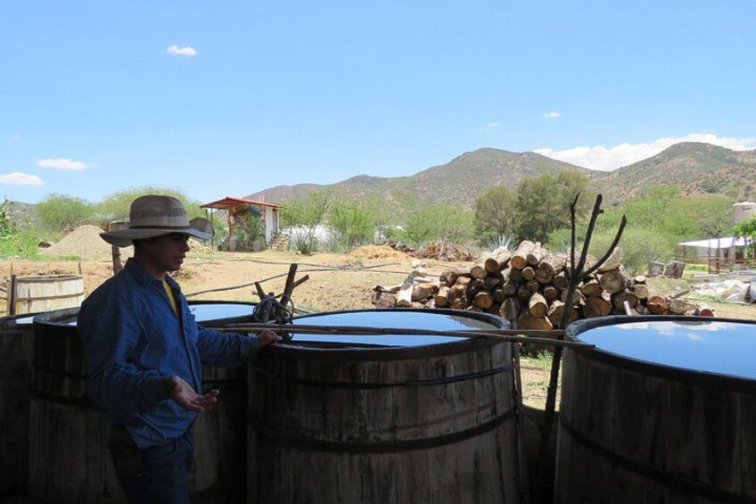 A man in a hat standing next to large wooden barrels in a rural setting with mountains in the background. 3