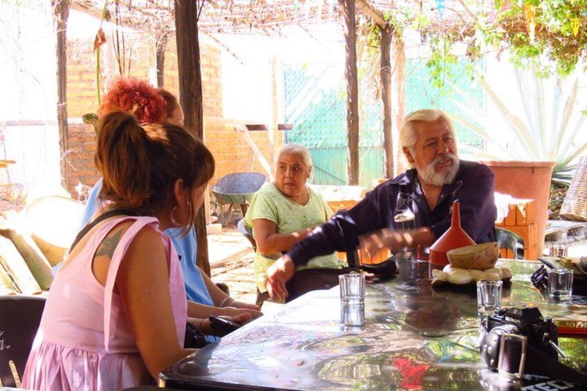 People sitting around a table with drinks and food, outdoors with trees and a fence in the background. 2