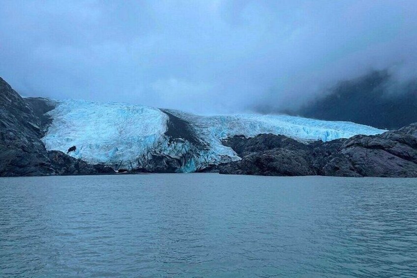 A glacier with a small waterfall, surrounded by rocky terrain and overcast skies. 2