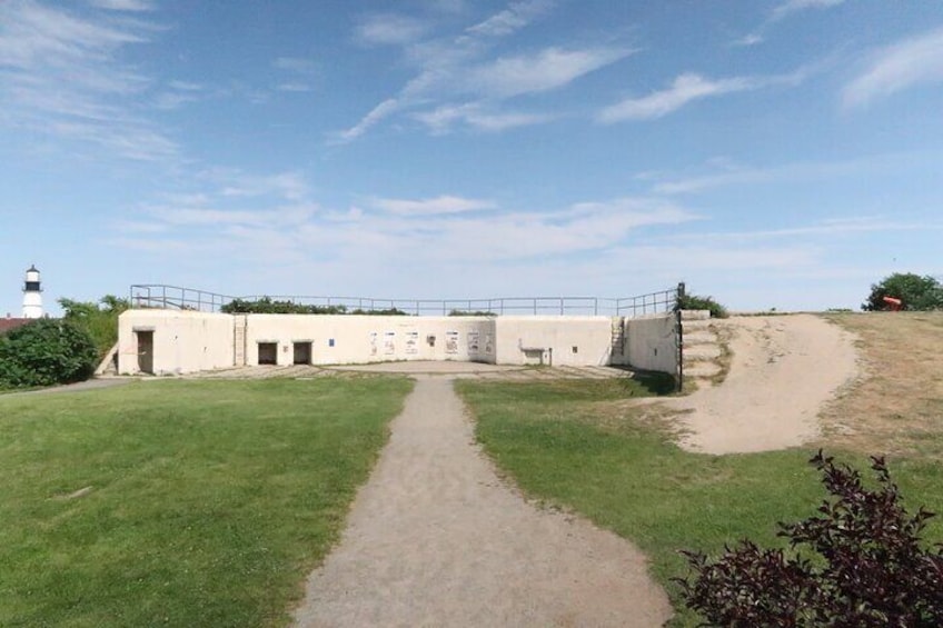 A coastal fortification with a lighthouse in the background, a dirt path leading to the entrance, and a grassy area. 2