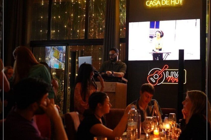 A dimly lit indoor venue with a stage, a large screen displaying a performer, and a neon sign reading 'CASA DE HOY.' 4