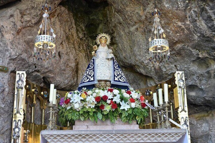 A religious statue with a blue and white robe, surrounded by flowers and candles, set against a rocky background. 2