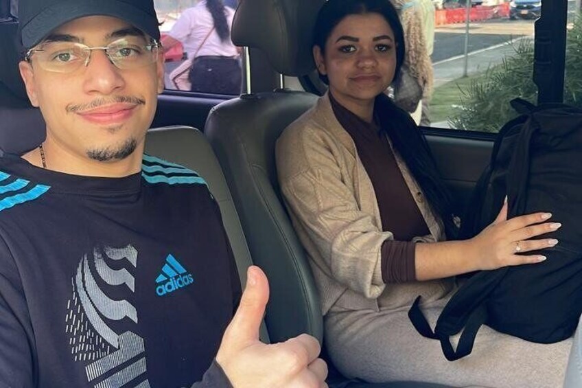 Two people in a vehicle with a black seat cover and a window showing a street scene. 5