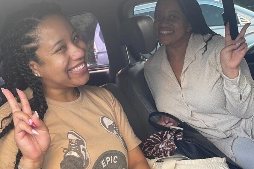 Two people in a car, one wearing a t-shirt with a shoe design and the other in a light-colored top. 2