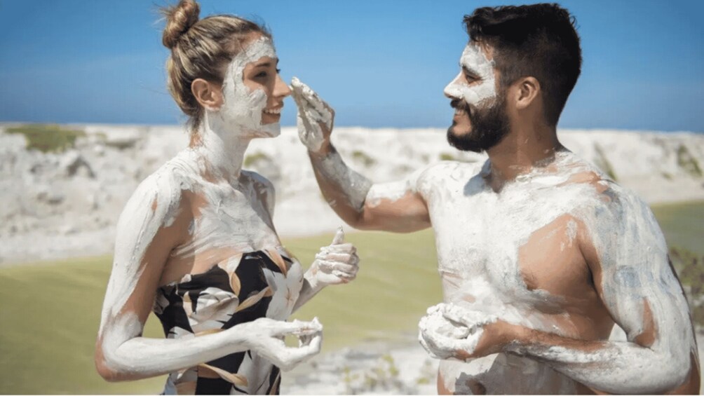 Two people covered in white substance, possibly mud or sand, at a beach with a clear sky and distant hills. 2