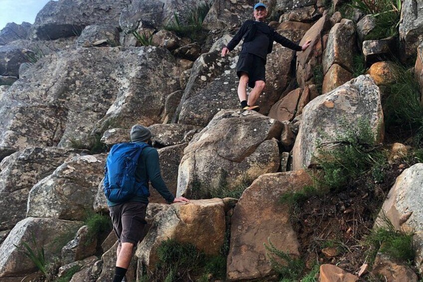 Two hikers ascending rocky terrain with large boulders and sparse vegetation. 5