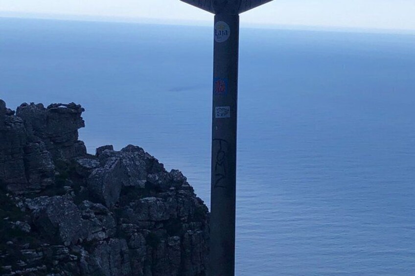 A rocky cliff with a metal pole in the foreground, a clear view of the ocean, and a distant horizon. 5
