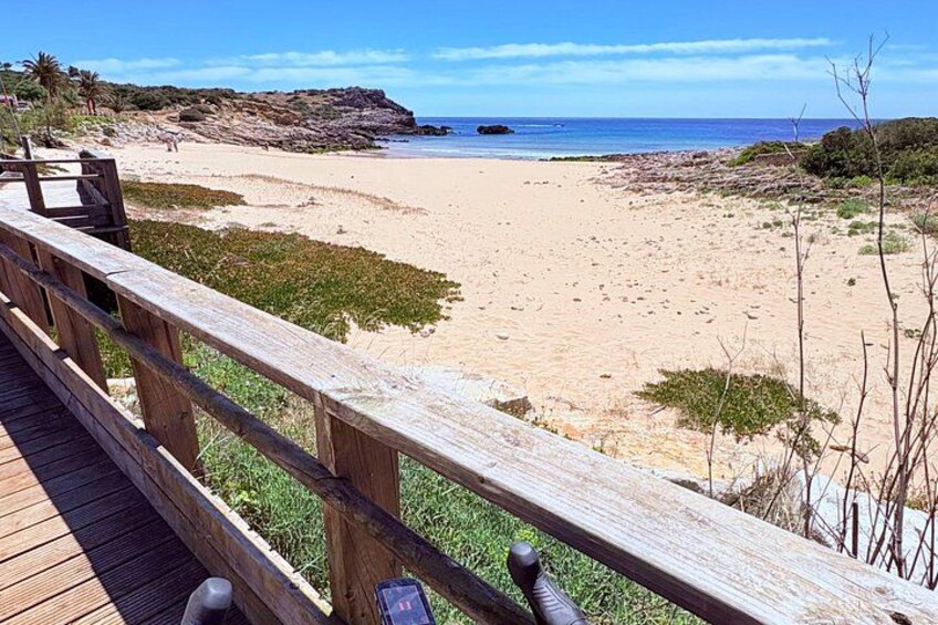 A wooden boardwalk leading to a sandy beach with sparse vegetation and a rocky outcrop in the distance. 2
