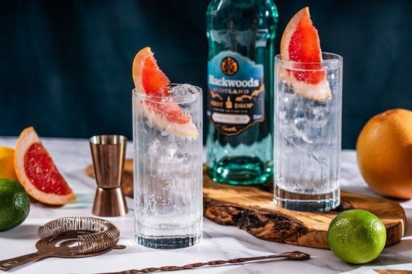 Two glasses of a clear beverage with ice cubes and a slice of grapefruit garnish, a bottle of Blackwoods Scotland Gin, and a wooden cutting board with a knife and a citrus fruit. 2