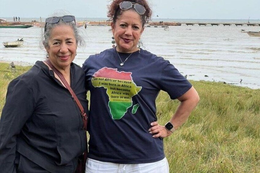 Two people standing by a body of water with a grassy area in the foreground and a pier in the background. 4