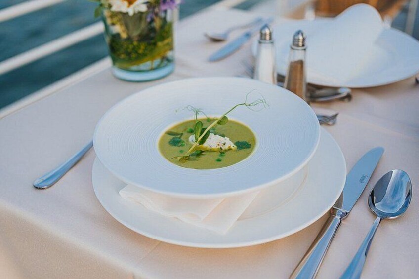 A white plate with a green soup garnished with herbs, silverware, and a salt shaker on a table. 2