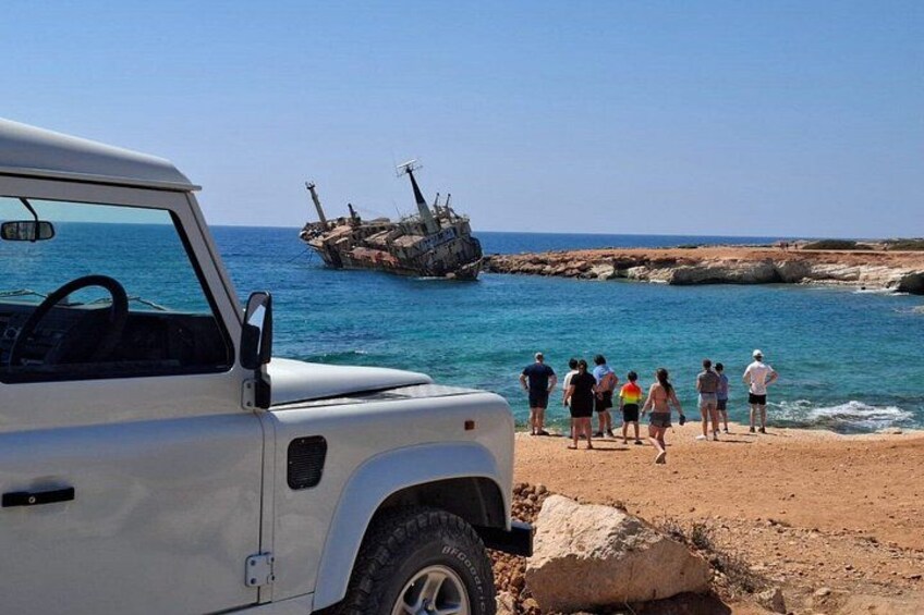 A white vehicle parked on a rocky beach with a group of people standing in the background. A large shipwreck is visible in the water. 5