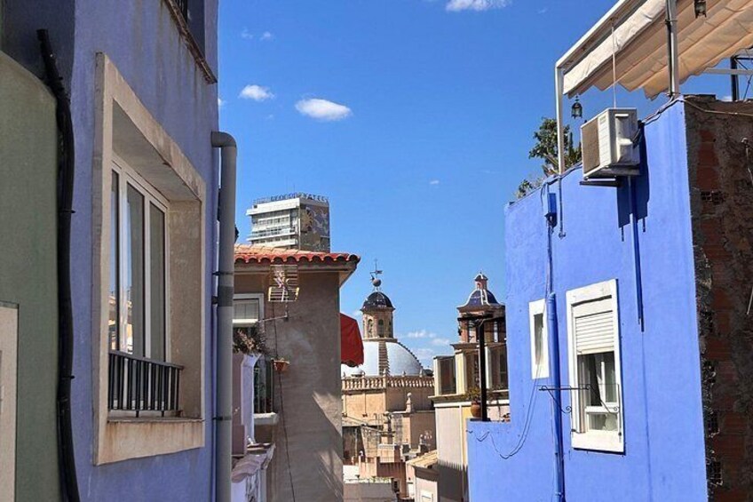 A narrow street with blue buildings, a church with a dome, and a building with a red roof. 2