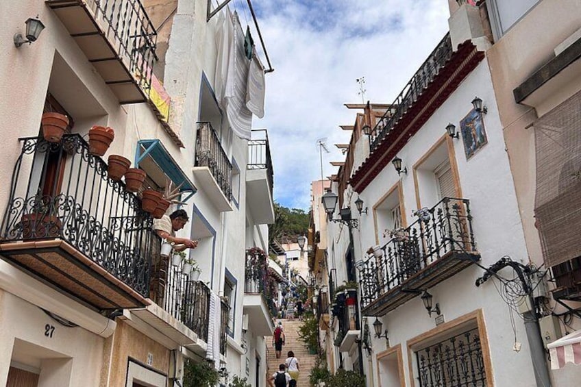 A narrow street flanked by buildings with balconies, some with potted plants, under a partly cloudy sky. 4