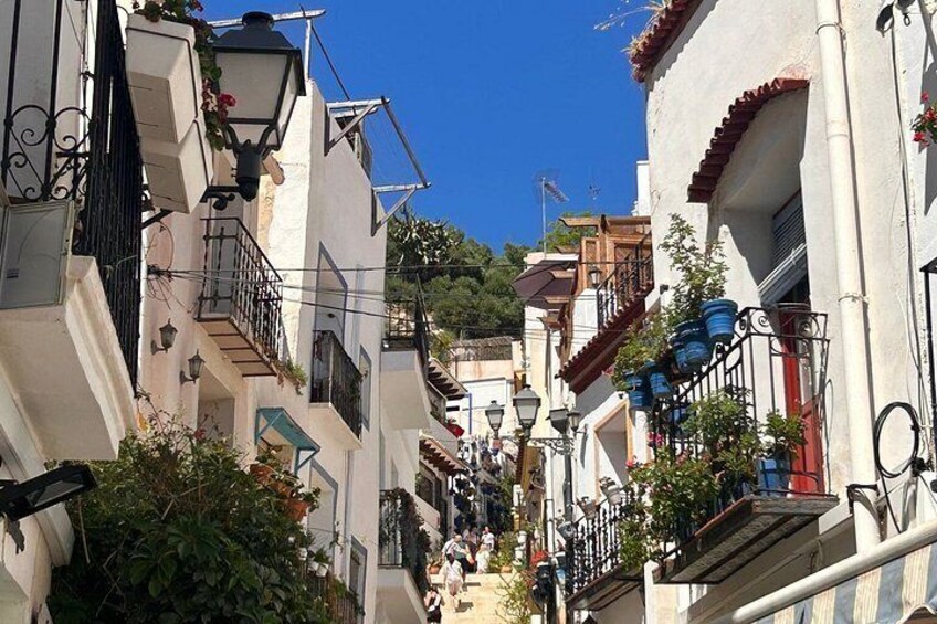A narrow street with buildings on both sides, a clear blue sky, and a few people walking. 5