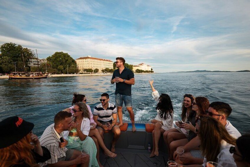 A group of people on a boat, some standing and some sitting, with a body of water and buildings in the background. 3