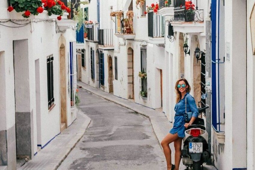 A narrow street with white buildings, a person in a blue dress, and a parked scooter. 2